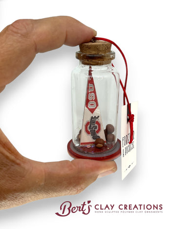 Hand holding a small bottle with a miniature flag inside, branded 'Bert's Clay Creations'.